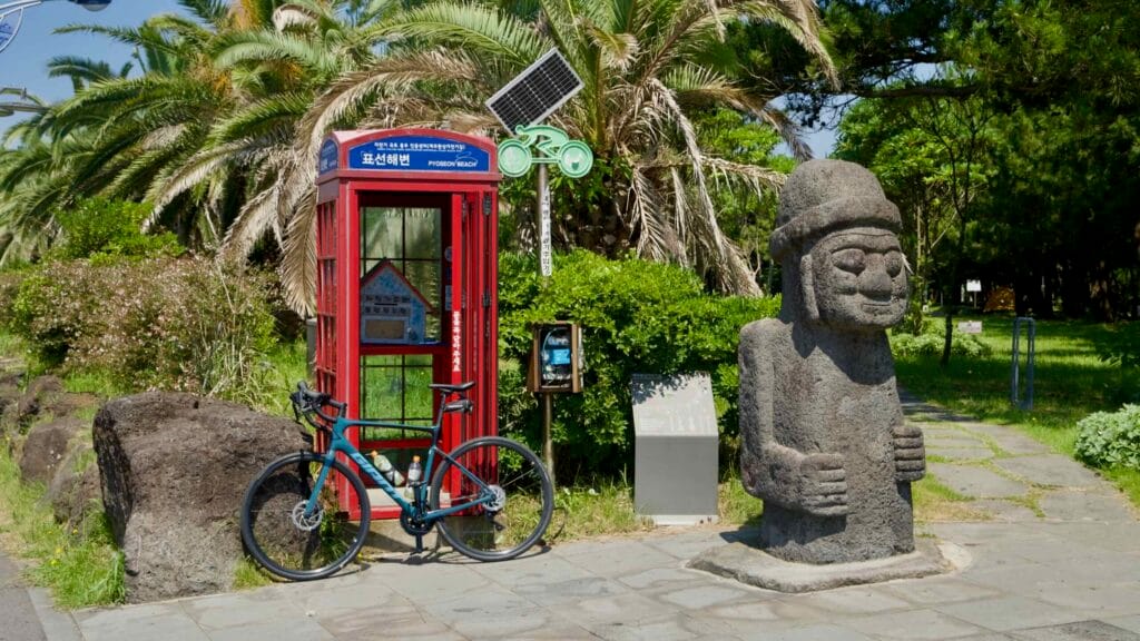 A dolhareubang statue stands by a red phone booth on the Pyoseon promenade with palms and pines behind.