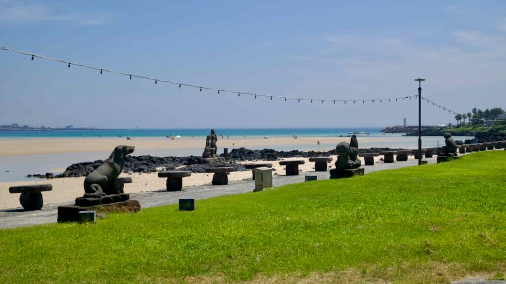 Stone stools and animal sculptures line the grassy promenade at Pyoseon Beach, facing the lagoon and open sea.