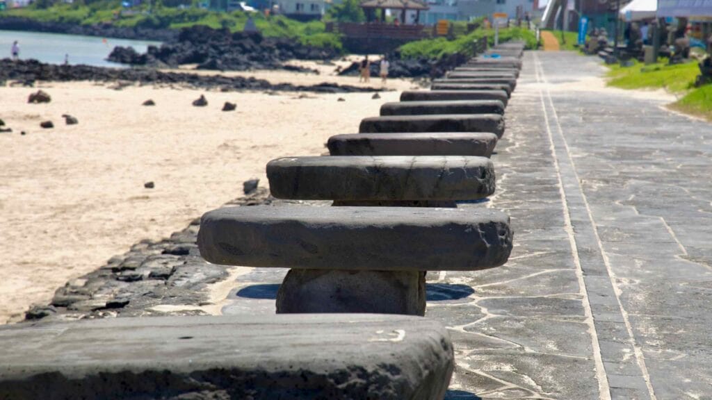 A row of circular basalt stools lines the Pyoseon Beach promenade above the sand with buildings.
