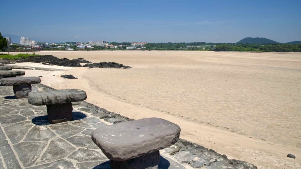 From basalt stools, the view opens across Pyoseon Beach flats toward village buildings and a low volcanic hill.