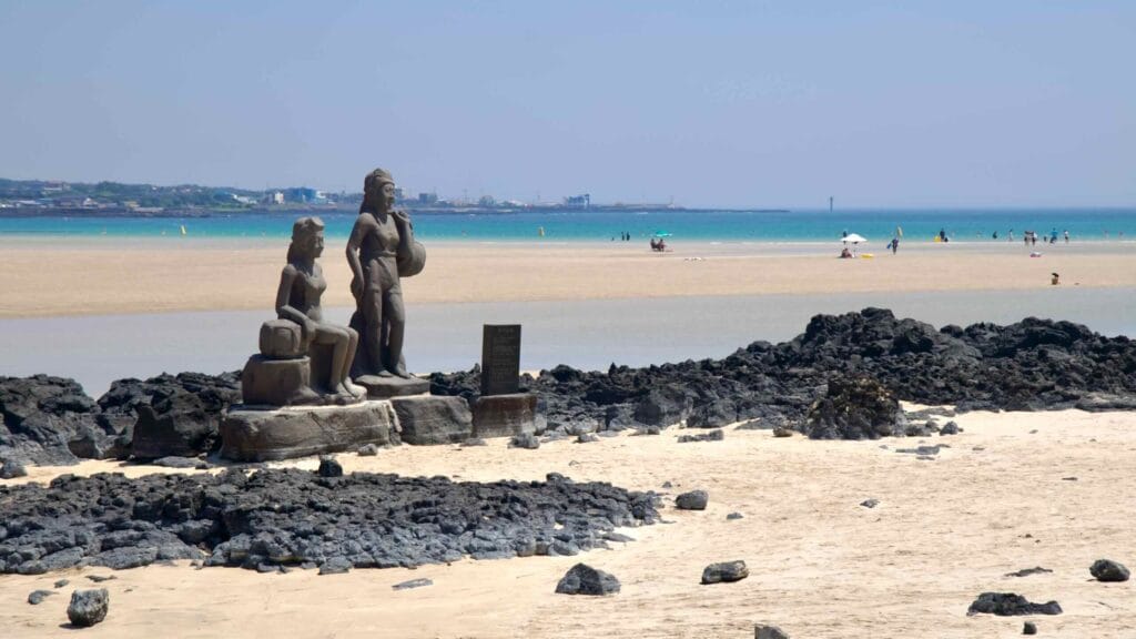 Statues of Jeju haenyeo divers rise from basalt by the Pyoseon lagoon as people relax on the sandbar.