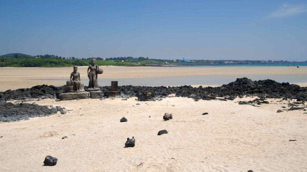 Haenyeo statues and lava rocks sit on Pyoseon Beach’s sand flat under a clear sky with lagoon.
