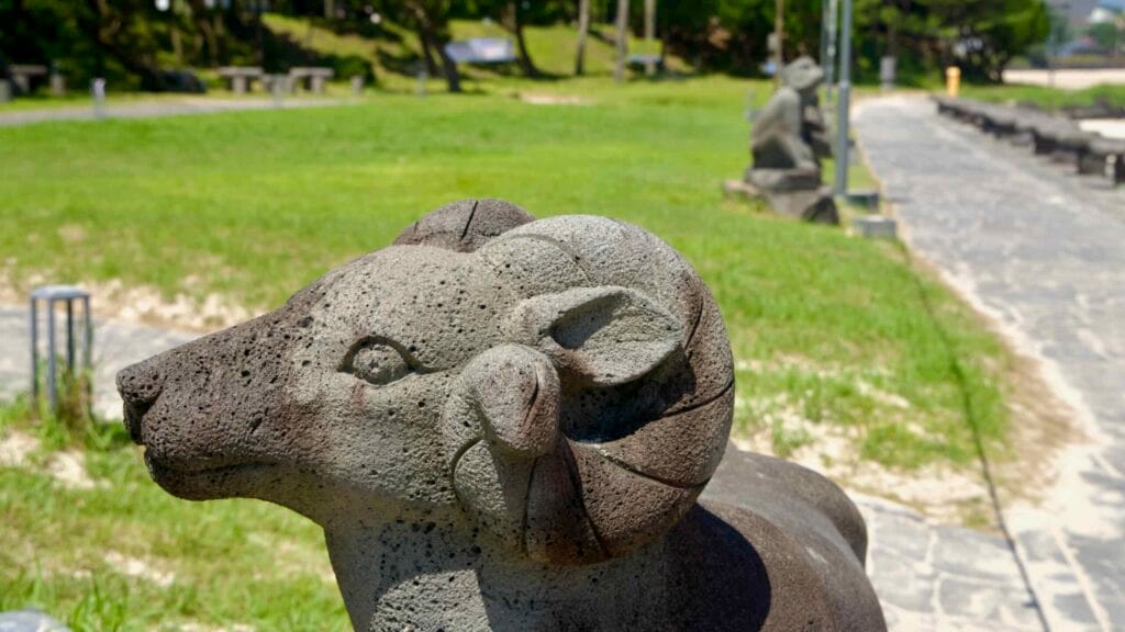A ram sculpture on the Pyoseon Beach walkway shows basalt texture and horns with more animal figures.