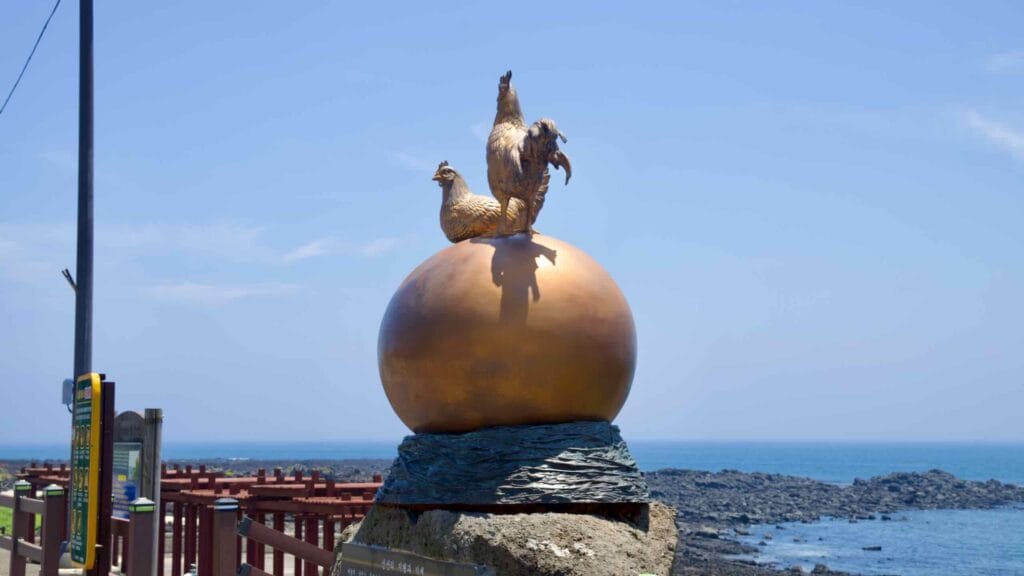 A rooster and hen sculpture crowns a seaside plinth above the lava shore on Jeju.
