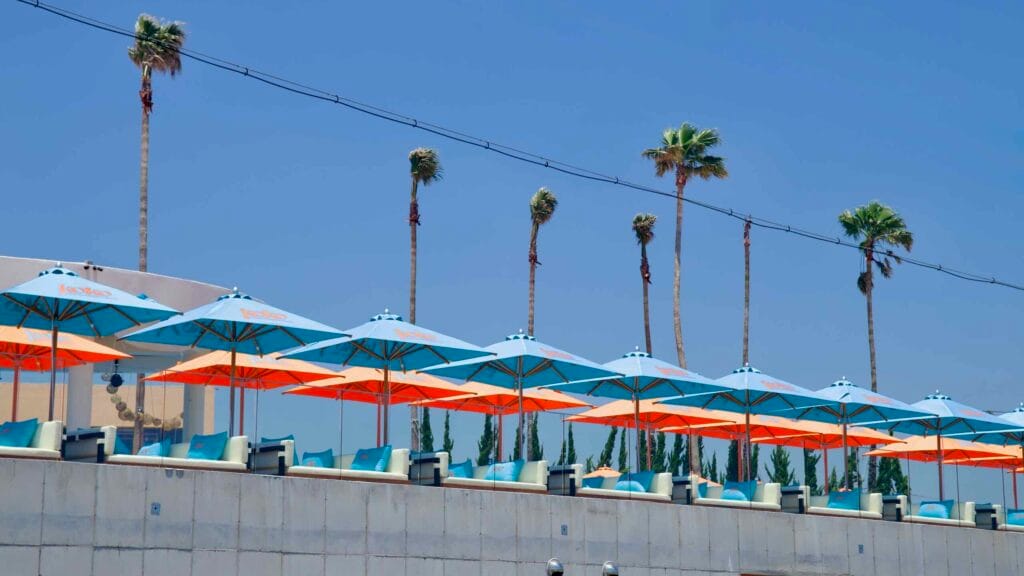 Turquoise and orange “koko” umbrellas line a rooftop terrace beneath tall seaside palms.