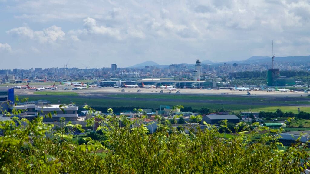 View of Jeju International Airport from Dodu Peak with jets, tower, and city skyline.
