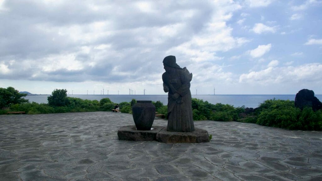 Stone haenyeo statue faces the sea near Handam with distant turbines on the horizon.