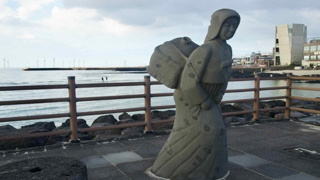 Stone haenyeo statue on the Hyeopjae promenade with turbines across the horizon.