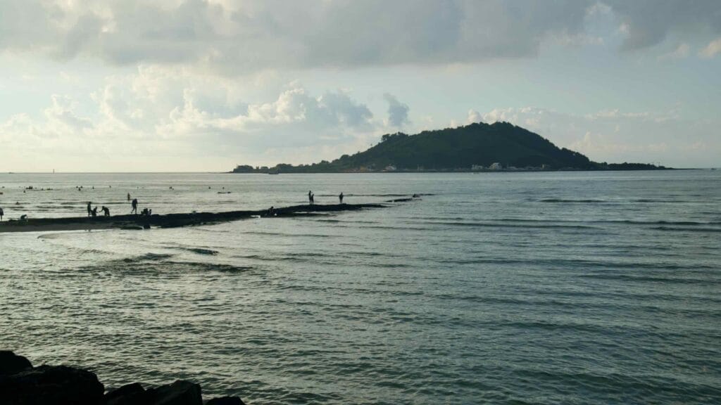 Visitors wade the tidal flats at Hyeopjae as Biyangdo rises offshore in soft evening light.