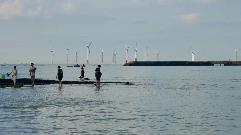 Visitors on a tidal ledge at Hyeopjae with a breakwater and offshore wind turbines beyond.