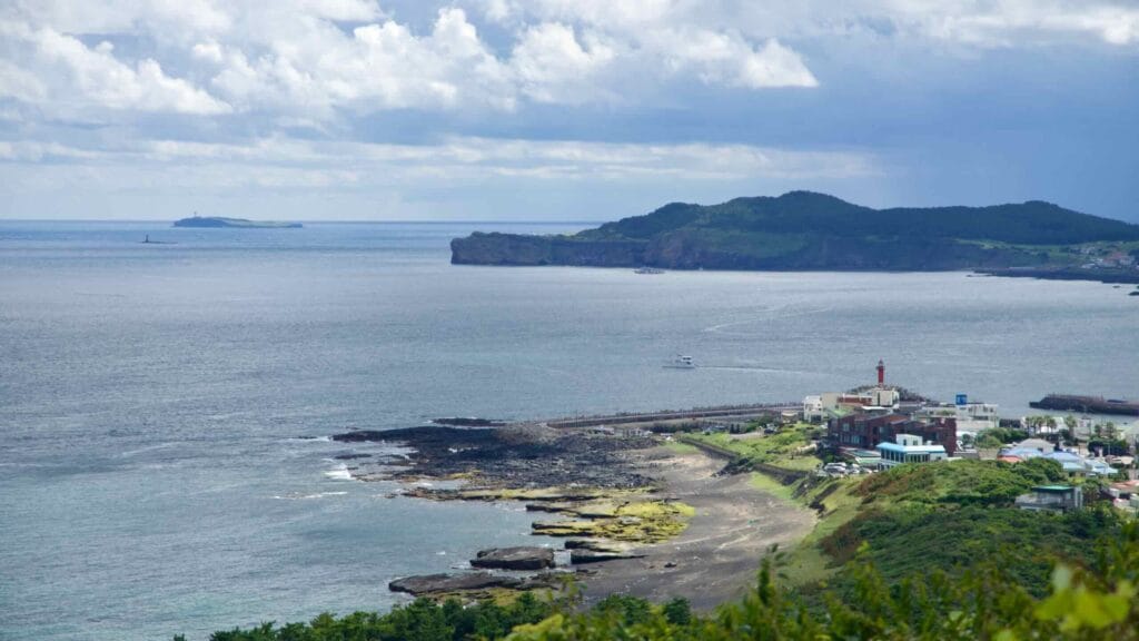 Distant view of Jeju’s Songak Mountain and a lighthouse from Sanbangsan, with boats and a rocky beach.
