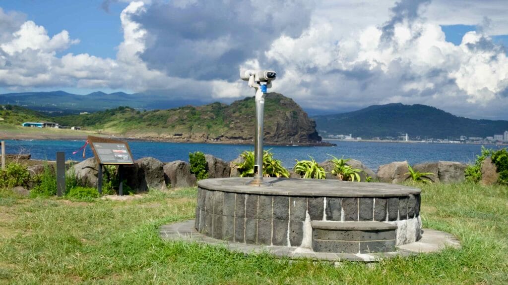 A viewing telescope on a stone platform faces the sea with Sanbangsan and Jeju’s dramatic coastline.