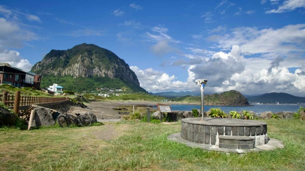 A wide view of Sanbangsan Mountain rises above Jeju’s coast, with a telescope stand and shoreline buildings.
