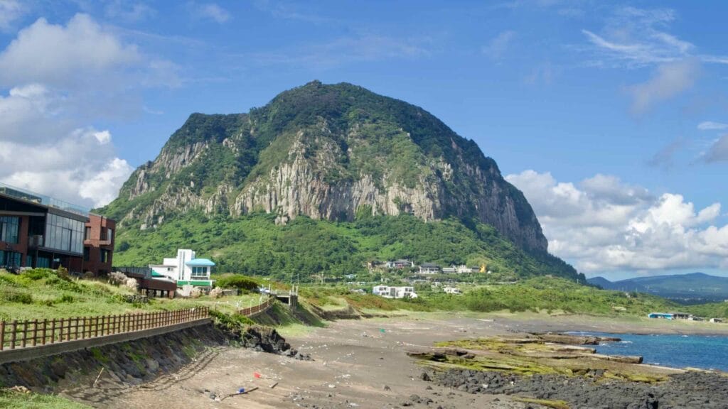 Sanbangsan Mountain dominates Jeju’s southern coast, with houses below and a black volcanic beach in view.