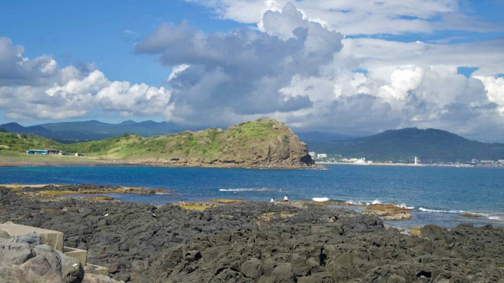 Yongmeori anchors Sagye Bay beneath towering clouds, with basalt benches and shallow tidal pools.