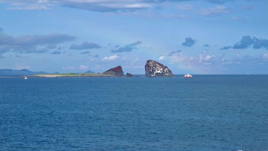 Twin Hyeongje Islets rise past a sandbar as a red ferry crosses the channel, seen from shore.