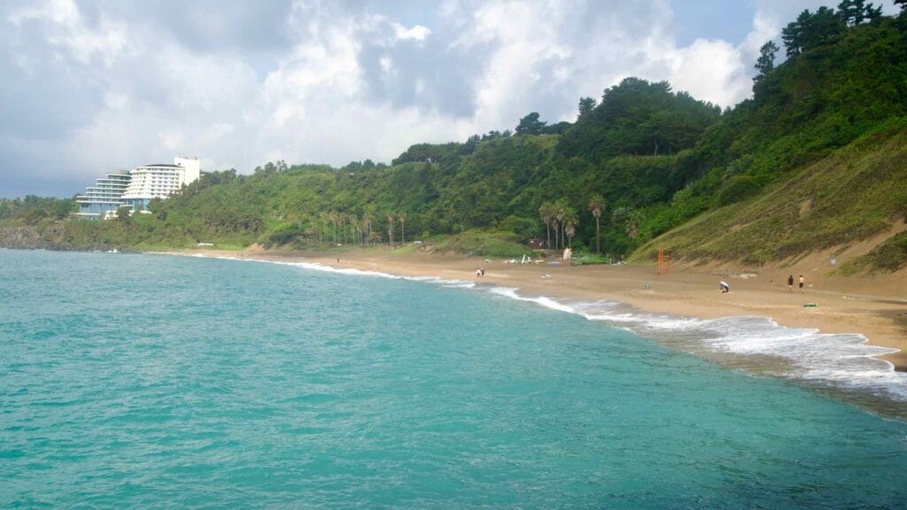 Turquoise water laps a wide arc of sand at Jungmun, with a resort and palms by green slopes.
