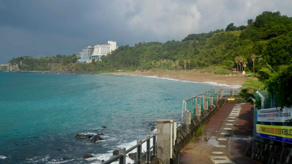 An elevated seaside walkway curves along basalt walls toward Jungmun Saekdal.