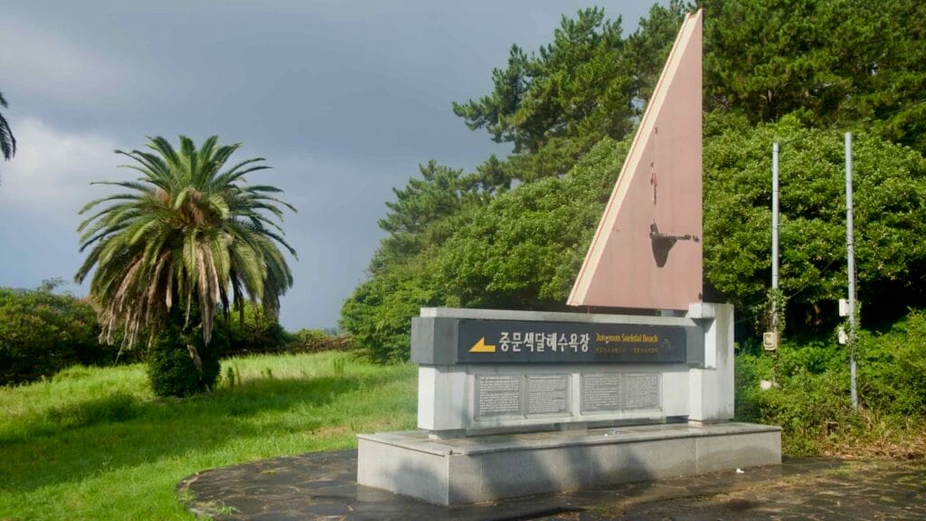 The Jungmun Saekdal Beach entrance monument, sail-shaped beside a palm, marks the way on a hot day.