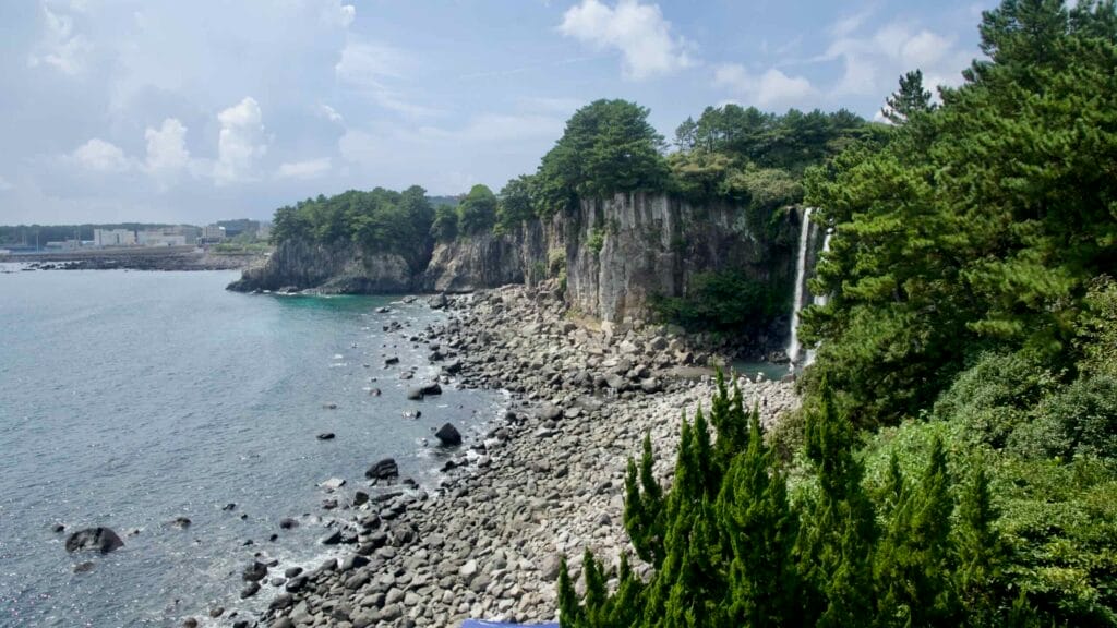 Jeongbang Falls spills from a forested cliff along a curving rocky coast.