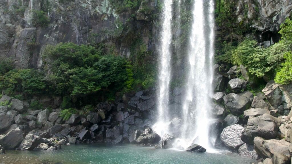 Lower cascades drop to a calm pool while sightseers wade at the edge of Jeongbang Falls.