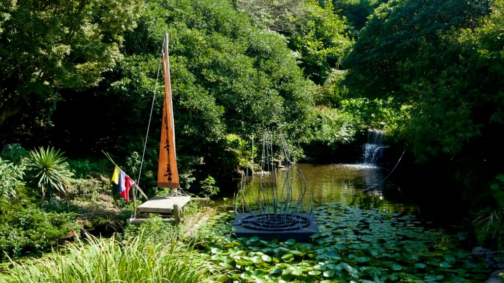 A pond at Jeju Folk Village holds water lilies, a low cascade, and a dock with a sail and sculpture among trees.