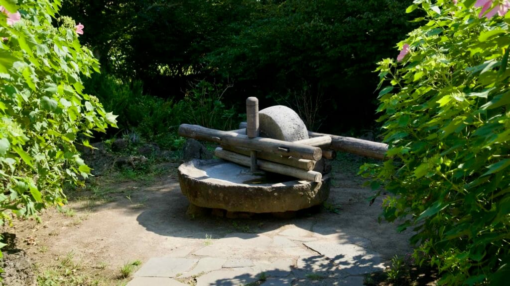 A rotary stone mill with wooden handle stands in a garden clearing at Jeju Folk Village for grinding grain.