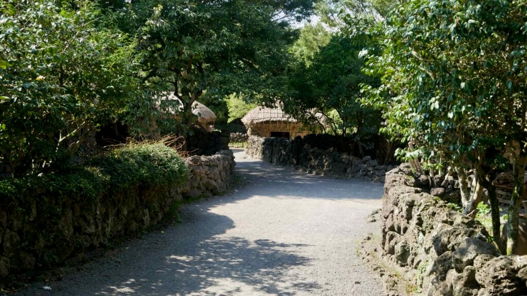 A quiet lane runs between basalt walls and camellia trees toward thatched cottages in Jeju Folk Village.