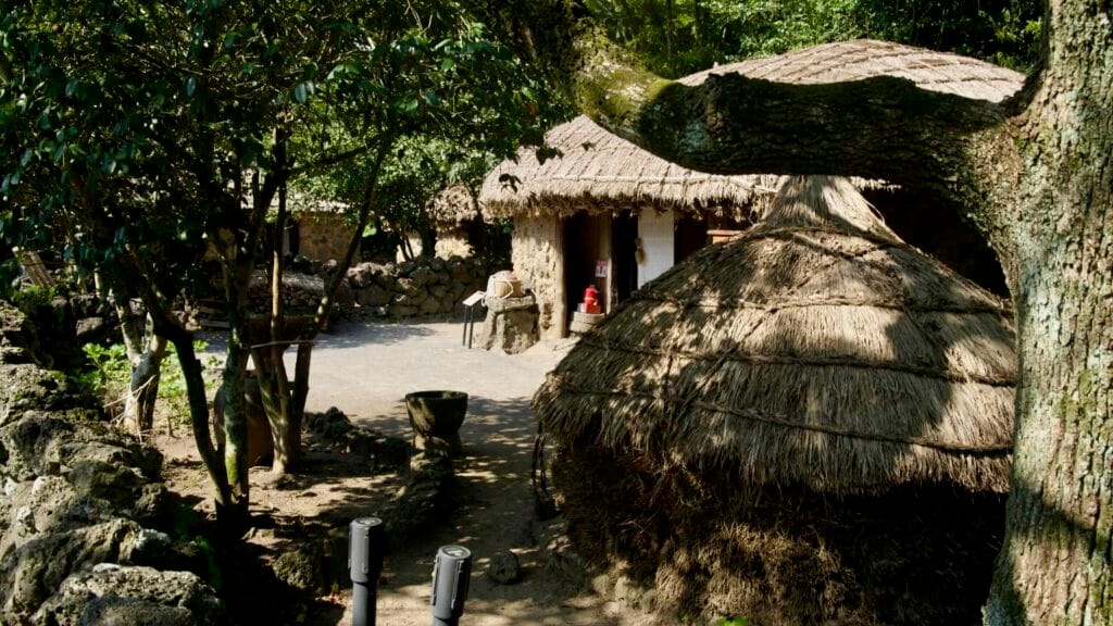 A courtyard at Jeju Folk Village shows a thatched storage stack and a cottage with household tools under shade.