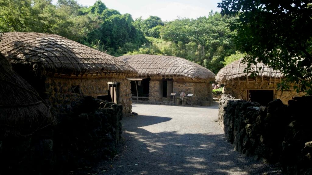 Low stone cottages with tight thatch roofs surround a yard at Jeju Folk Village, framed by walls and forested slopes.