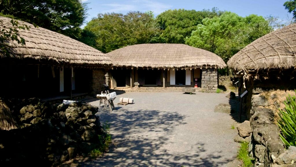 A courtyard enclosed by thatched houses at Jeju Folk Village shows benches and tools.