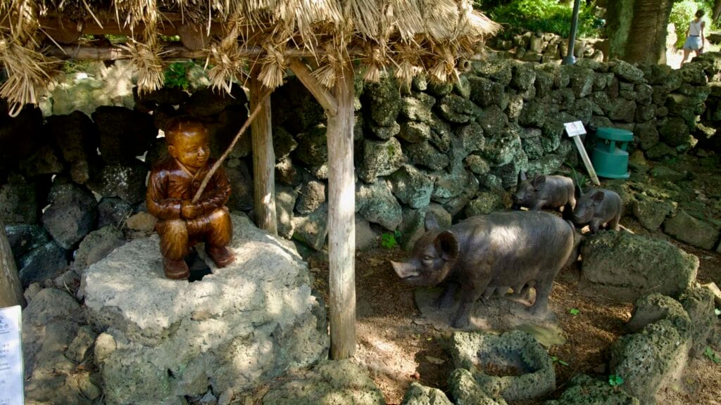 Carved figures of a boy and sow with piglets sit in a lava-stone pen at Jeju Folk Village, showing farm life.