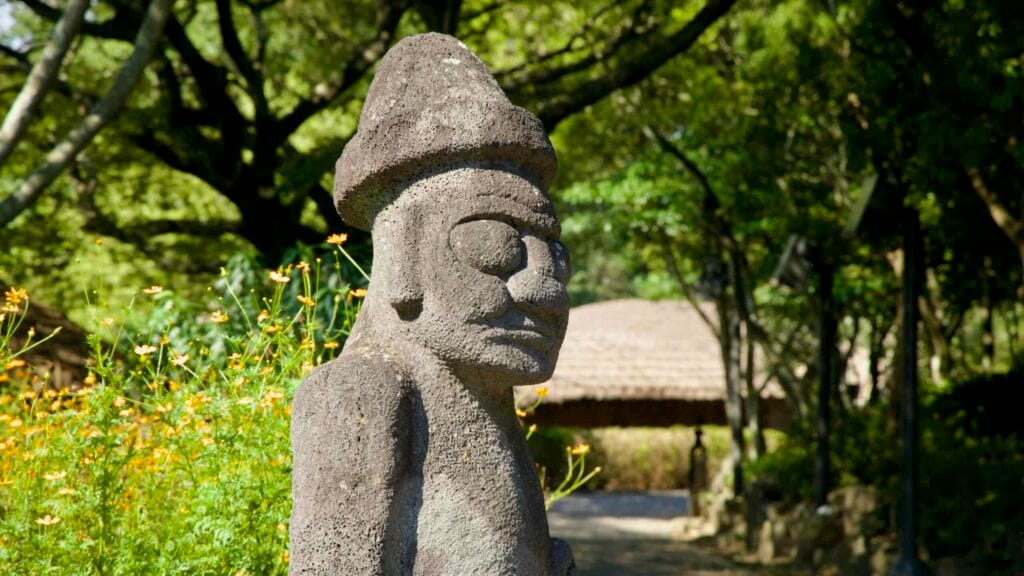 A dolharubang statue carved from basalt stands by wildflowers at Jeju Folk Village.