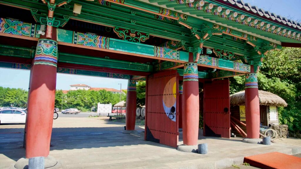 The painted gate at Jeju Folk Village shows dancheong patterns and red doors, marking the entrance from parking.