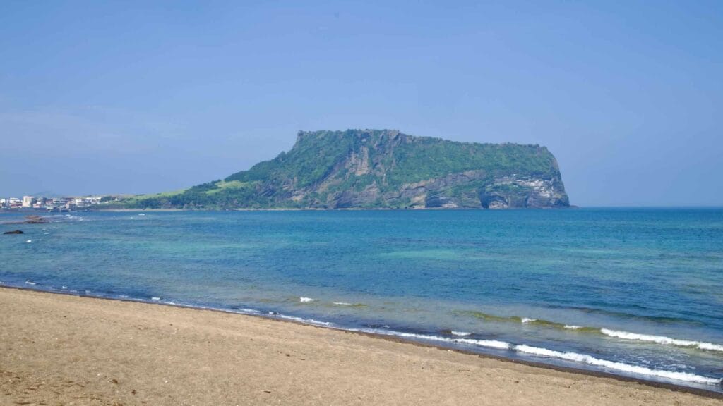 From the sandy shore, Seongsan Ilchulbong’s crater rim and layered cliffs rise across calm blue water.