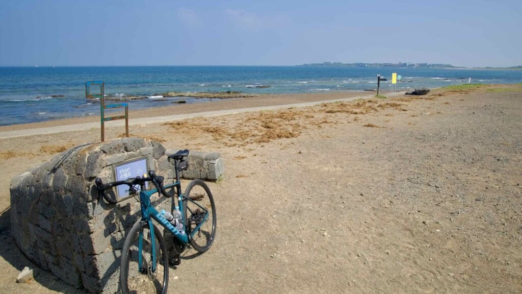 At the beachside stamp stone on the Jeju cycling path, a road bike rests above volcanic sand.