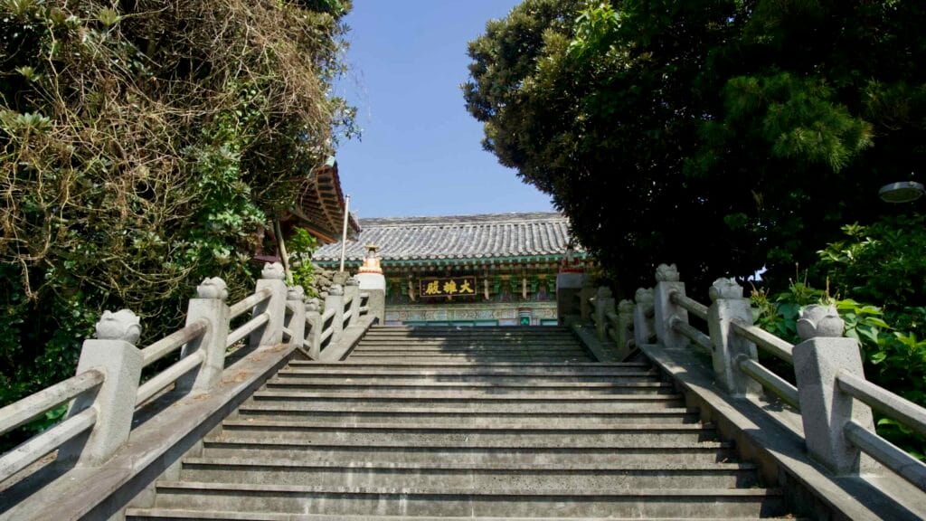 Stone steps lined with sculpted balustrades lead to a traditional temple hall with dancheong patterns beneath pine shade.