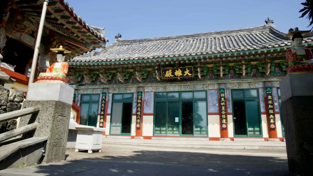 Colorful guardian statues flank the steps to a Daeungjeon main hall near Seongsan, with bright dancheong beams and tiled roof…