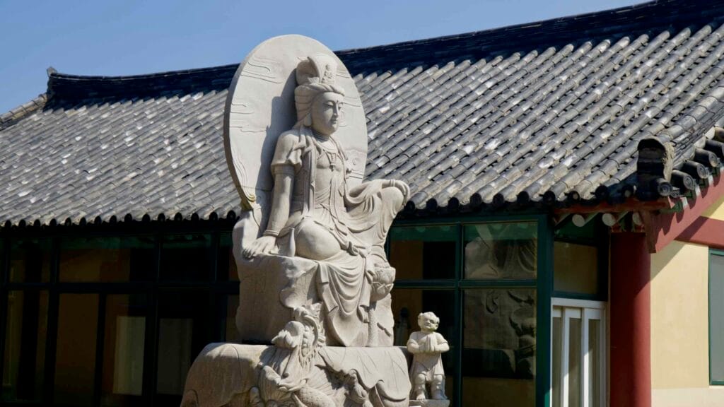 A carved bodhisattva statue with halo, dragon, and wave motifs stands in the temple yard near Seongsan, its pale stone glowin…