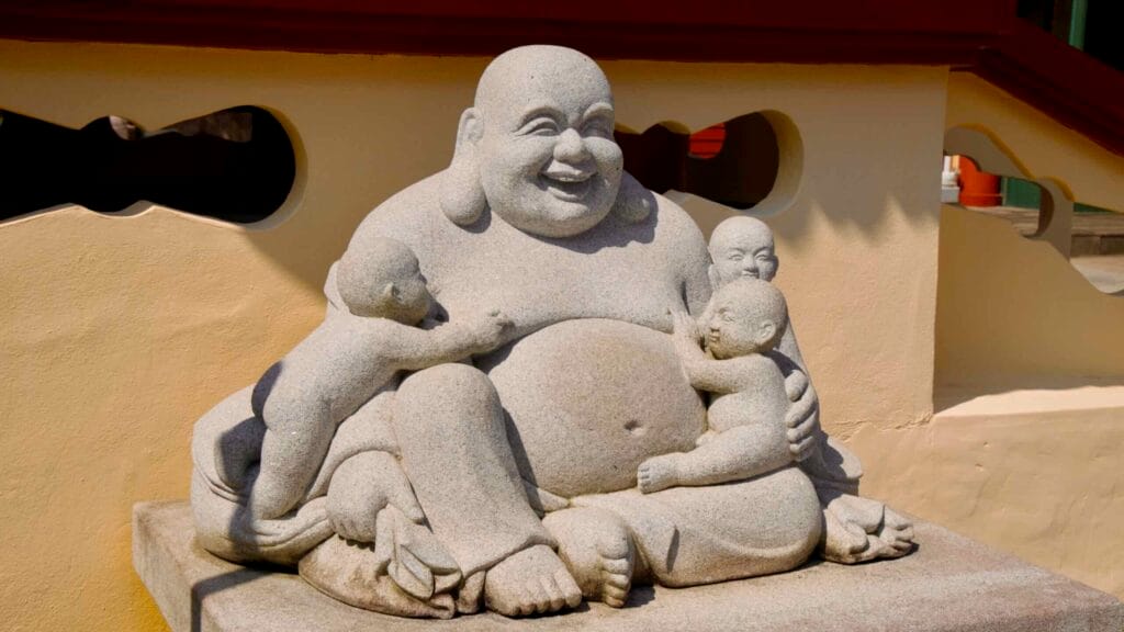 A granite statue of the Laughing Buddha surrounded by playful children sits along a temple.