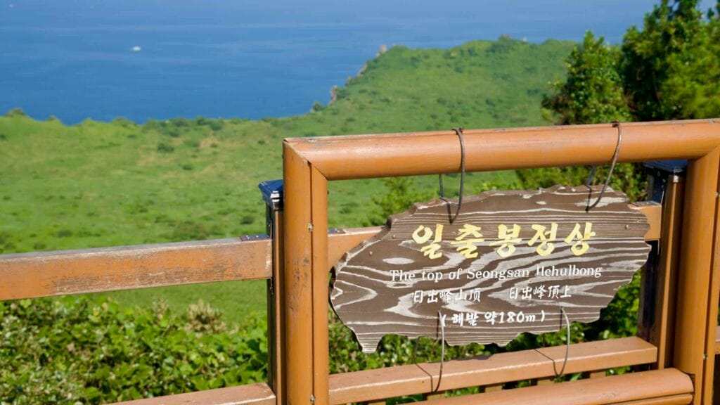 A summit sign reading “The top of Seongsan Ilchulbong” hangs on a wooden fence.