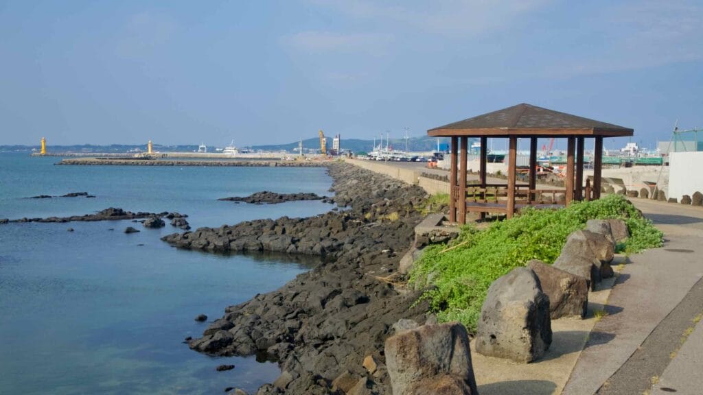 A seaside walkway leads past basalt rocks toward Seongsan’s small harbor.