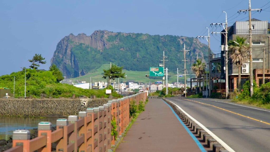A waterfront path runs beside the coastal road with Seongsan Ilchulbong visible beyond.