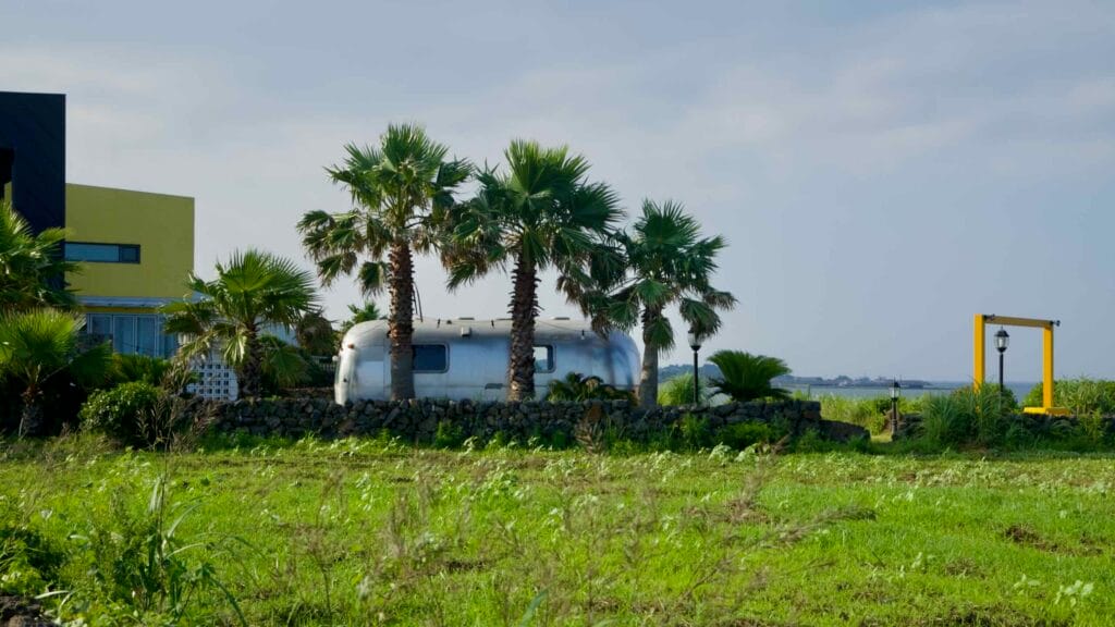 A retro silver trailer rests among palm trees behind a low stone wall near the coast.