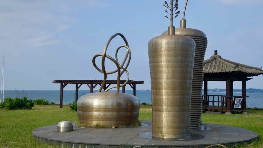 A polished metal sculpture stands beside a pavilion on the Dumun Port waterfront lawn.