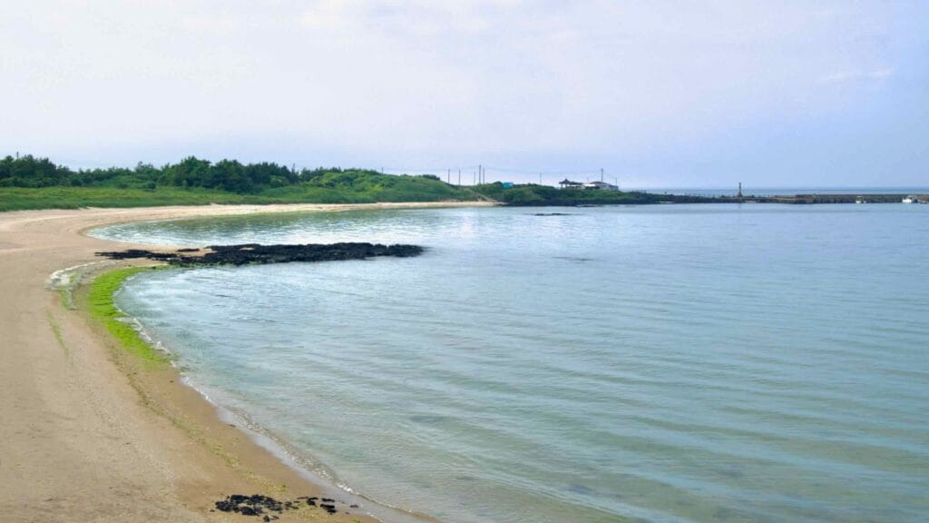 The curving shore of Hado Bay leads toward a stone breakwater and small lighthouse.