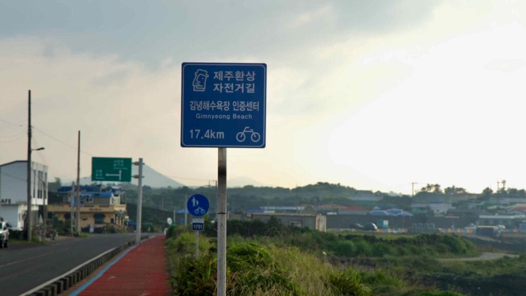 A blue bikeway sign points toward Gimnyeong Beach along Hado’s scenic coast.