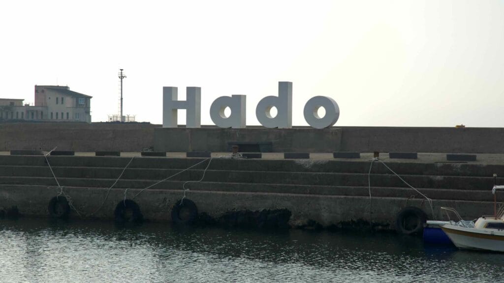 Large white “Hado” letters stand on the harbor jetty with an angler nearby.