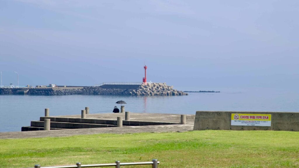 A red lighthouse marks the breakwater at Pyeongdae Port beyond calm water and tetrapods.