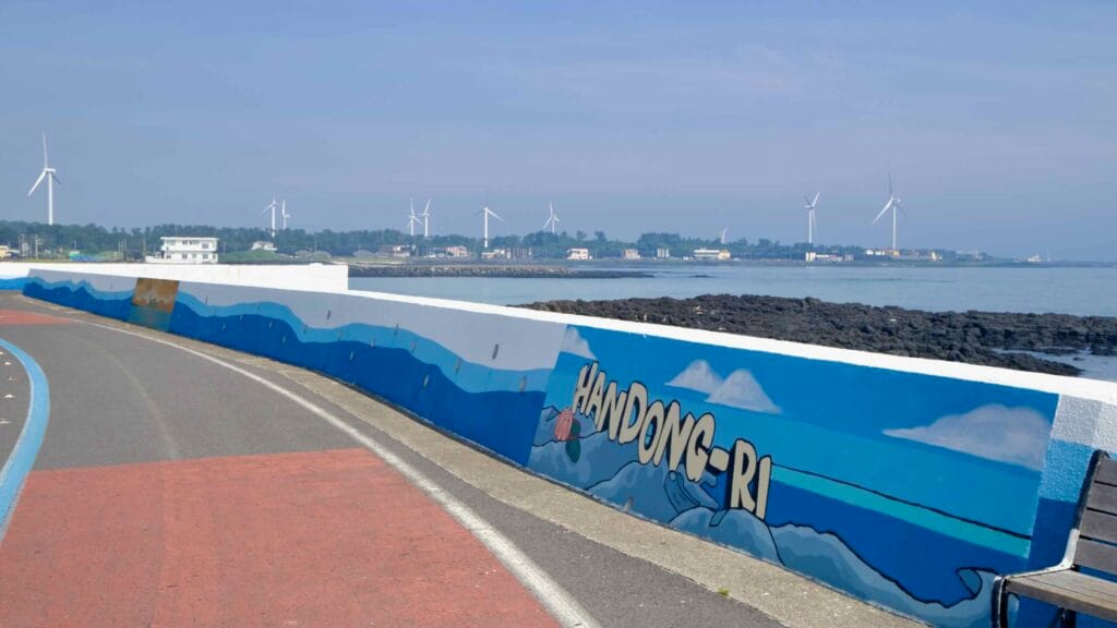 A Handong-ri seawall mural lines the coastal bike lane with wind turbines beyond.